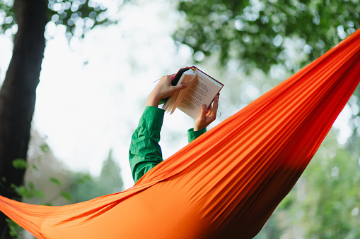 Close-up view of a woman's hands delicately grasping a book while engrossed in reading on a hammock in the park, capturing the intimacy of the moment