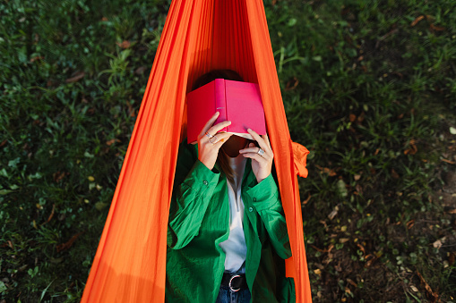 Woman lying in a hammock with a book resting on her face, portraying a whimsical and relaxing scene of leisurely reading outdoors