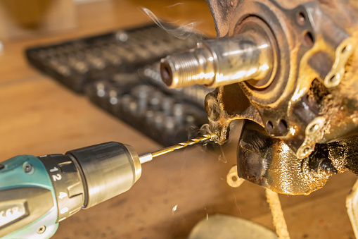 The car mechanic drills a thread on the rear wheel hub bearing. Smoke produced. Auto service. Opened tools box in the background. Yellow light.