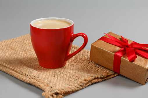 Red cup of black coffee and a gift box tied with red ribbon on the sackcloth bag. Greeting card concept.