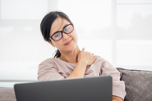 Mature middle age asian woman feeling pain in neck and shoulder after working on computer laptop for a long time in living room at home . She stretches to relax her muscles.Office syndrome concept.