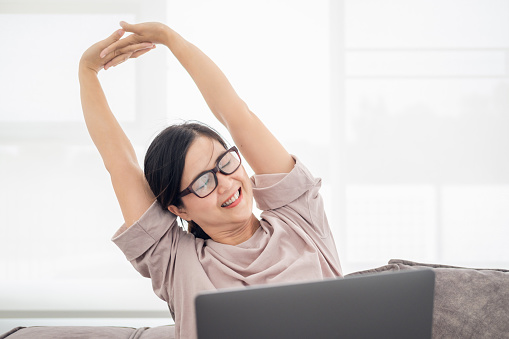 Mature middle age asian woman feeling pain in neck and shoulder after working on computer laptop for a long time in living room at home . She stretches to relax her muscles.Office syndrome concept.