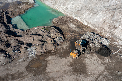 A large, yellow dump truck unloads gravel in gravel pit next to a small green lake, aerial view.