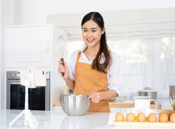 istock baker online courses. food preparation and culinary training class concept. smiling woman chef prepare ingredients in the kitchen and shooting video of herself using mobile phone on a tripod. 1709494371