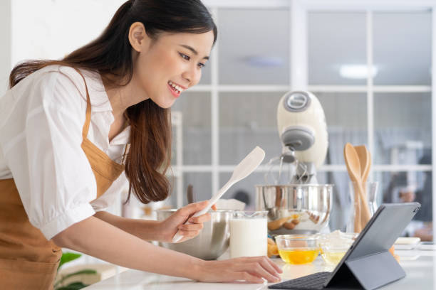 istock Young asian houes wife learn cooking online. A young woman is watching cooking tutorial video in the kitchen.Reading recipe while making cookies Following Recipe On Digital Tablet. 1709496550