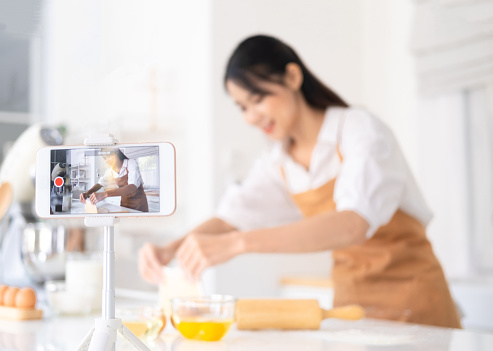 Portrait of concentrated young woman food vlogger showing process of baking in live stream, standing in front of smart phone on tripod, broadcasting. Indoor studio shot on kitchen background.