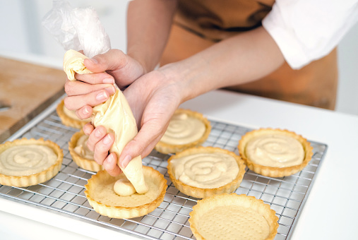 close up hands of Small business entrepreneur asian woman bakery shop making fruit tart. Female Baker Using Piping Bag to Decorate fruit tart with cream cheese on white kitchen counter.