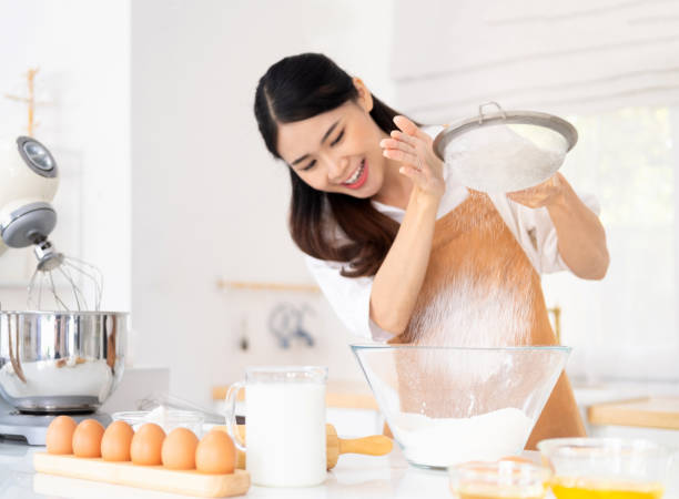 woman baking at home. young woman wearing light brown apron in the modern kitchen have fun sifting bread flour, preparing different ingredients before mixing, to make homemade bread. - chef cookie dishware domestic kitchen imagens e fotografias de stock