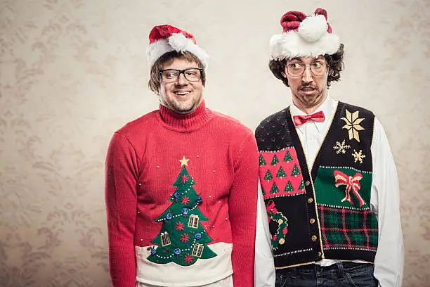 Two goofy looking men in ugly looking Christmas cardigans and sweaters (complete with matching red bow tie and a classy mustache) stand looking awkward for a holiday photo.  Damask style vintage wall paper in the background.  Horizontal with copy space.