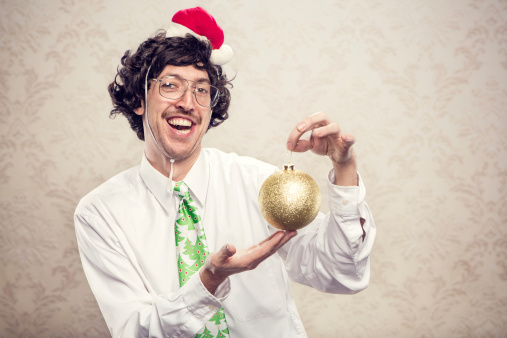 An office nerd in a white collared shirt with a Christmas tree tie shows his holiday spirit, holding up a giant golden bauble with a goofy look on his face.  He has big glasses and a mustache.   A miniature Santa hat is on his head.  Horizontal with copy space on wallpaper background.