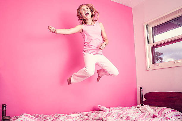 Girl Jumping on Bed in Pink Room An elementary aged girl plays air guitar and screams and sings in her pink colored room as she jumps on her bed. Horizontal with copy space. pink bed stock pictures, royalty-free photos & images