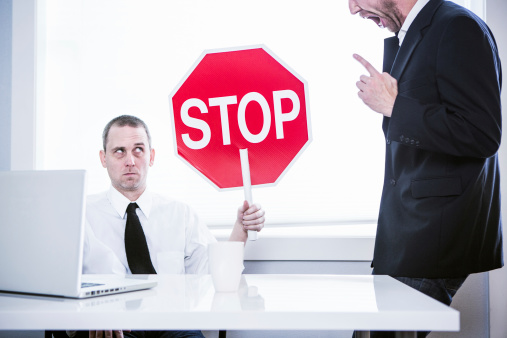 A business man sits at his office desk in front of his computer, while his CEO yells loudly at him.  He holds up a stop sign with a funny look on his face, ready to be finished with his job.  Horizontal.