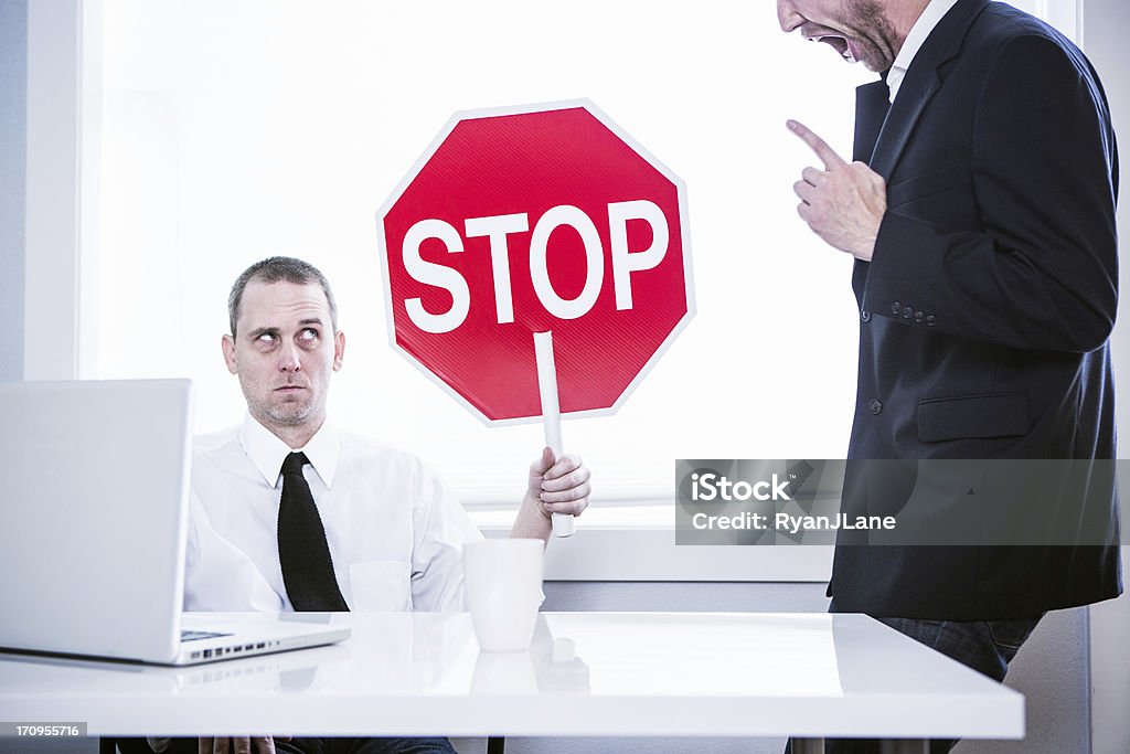Abusive Office Manager A business man sits at his office desk in front of his computer, while his CEO yells loudly at him.  He holds up a stop sign with a funny look on his face, ready to be finished with his job.  Horizontal. Conflict Stock Photo