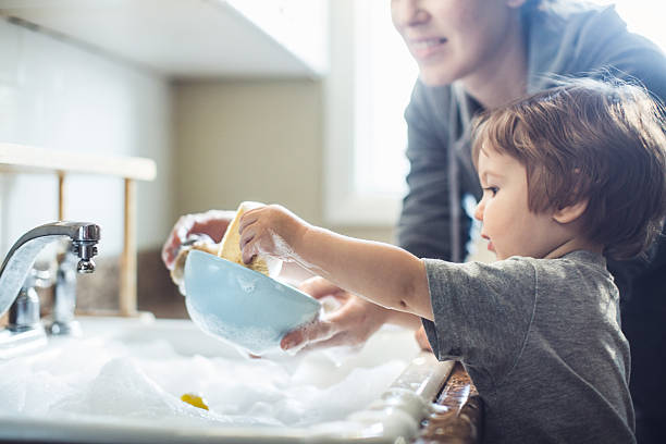 Baby Dish Washing A cute toddler aged boy wearing a blue diaper and tshirt helps out with with washing the dishes, standing on a chair to be able to reach the sink full of soapy bubbles and dishes. His mother smiles behind him as she supervises his sensory perception experience. Bright sunlight comes in through the window behind him, lighting the sparse modern kitchen. wash dishes stock pictures, royalty-free photos & images