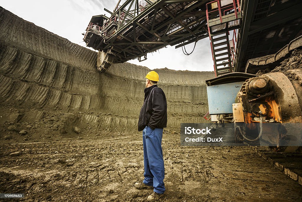 Heavy mining industry worker Coal mine worker with a helmet on his head standing in front of huge drill machine and looking at it. Rear view. Mining - Natural Resources Stock Photo