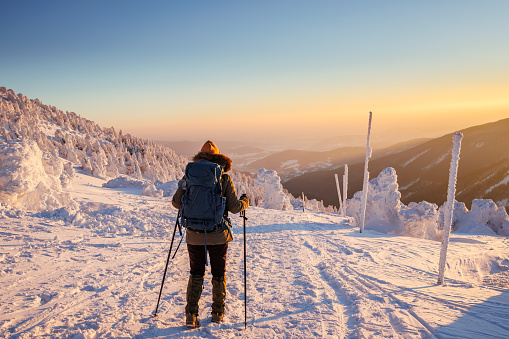Hiking in snow at winter mountain during sunset. Woman with backpack and nordic walking poles trekking in cold weather. Sports and outdoors seasonal activity
