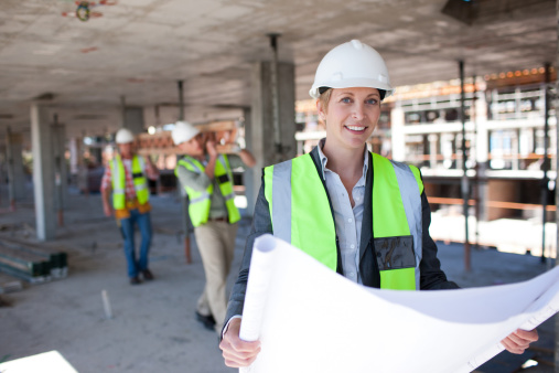 Portrait of woman engineer at building site looking at camera with copy space. Mature construction manager standing in yellow safety vest and yellow hardhat with crossed arms. Successful confident architect at construction site with team discussing in background.