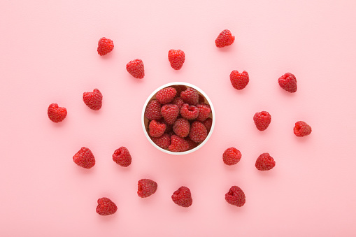 Fresh red raspberries in white bowl on light pink table background. Pastel color. Healthy food. Closeup. Top down view.