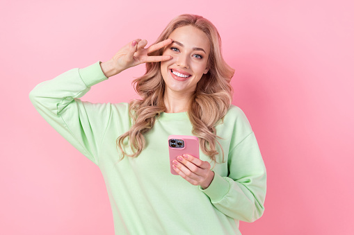 Portrait of young girlfriend showing v sign over her face blogging influencer wear green trendy pullover isolated on pink color background.