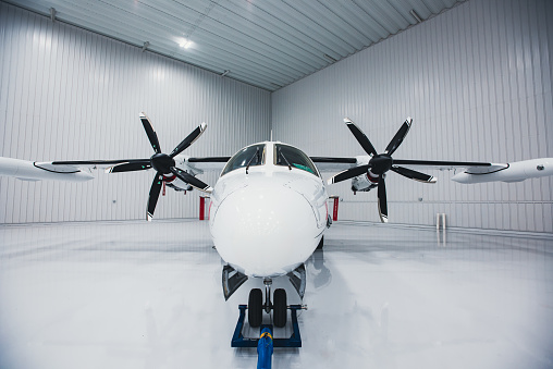 A twin-engine turbo prop aircraft parked in a hangar. The tow bar is attached to the front undercarriage.