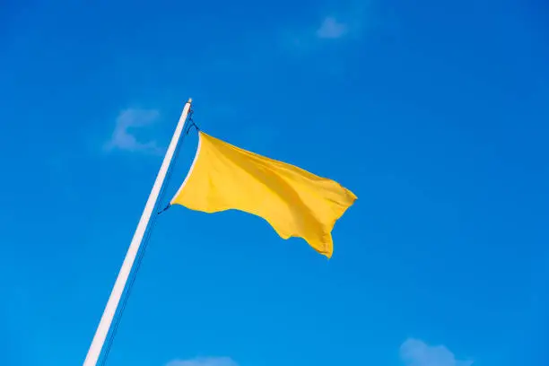 Yellow fabric flag waving to the wind with the background of a clear sky with white clouds.
