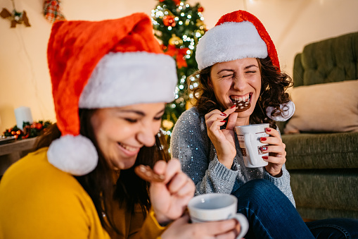 Two female friends eating Christmas cookies and drinking hot chocolate in a decorated home.