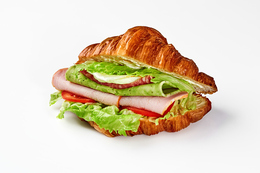 Appetizing sandwich from puff croissant with thin slices on ham, salami, spicy green cheese, fried egg, ripe tomatoes and fresh lettuce leaves on white background. French style breakfast