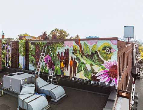 Two Female artists  painting roof top mural. They are dressed in casual work clothes. Exterior of old building in the city of Toronto, Canada.Drone point of view.