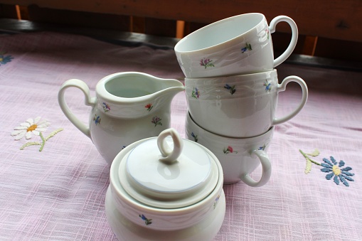 Parts of a romantic flowered coffee service with cups, milk jug and sugar bowl. The linen tablecloth has hand-embroidered flowers.