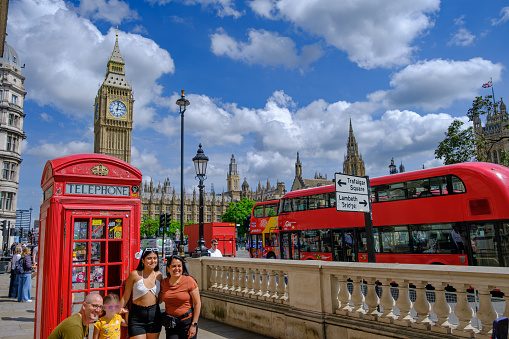 London, England - June 19, 2023: Tourist family taking a photo in fornt of a red traditional telephone booth in London with Big Ben Clock Tower and the Parliament in the background.