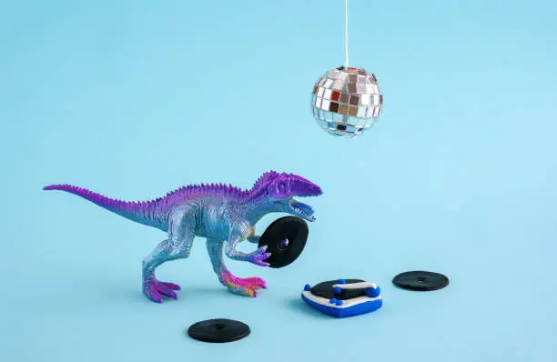 Photo of Cute blue and violet dinosaur toy holding listen vinyl on vinyl record player on blue background.