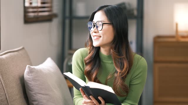 Smiling asian woman reading book at home, relaxing on a couch