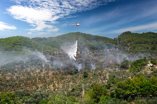 Firefighting helicopter with a bucket suspended on a cable, collecting water from a mountain lake to extinguish the flame in a forest