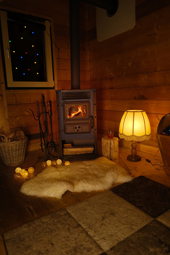 cozy wooden house with New Year's garlands, lights, warm light of lamp with lampshade, fire burning in fireplace, white sheepskin on floor, magical festive Christmas Anticipation, Dark Room Ambiance