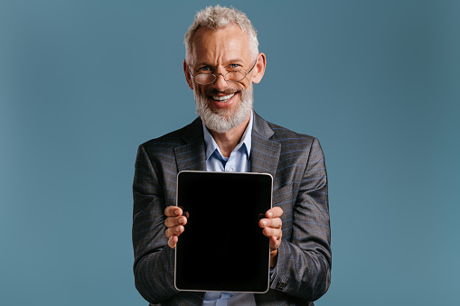 Happy mature businessman showing touchscreen of his digital tablet against colored background
