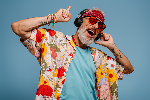 Joyful senior man in funky shirt and headphones listening to the music and dancing on blue background