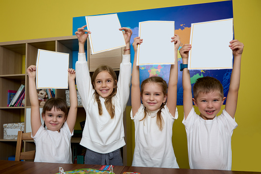 Satisfied children stand against background of yellow wall in office raised their hands up with white sheets of paper. Schoolchildren stand with paper in their hands raised at desk in the classroom