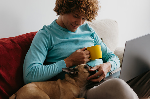 Young man sitting on the comfortable couch at home and petting dog while working from home