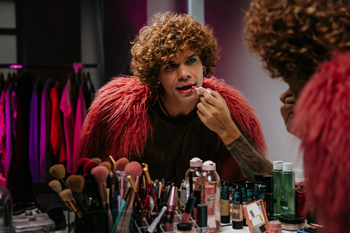 Confident young gay man in stage costume applying make-up while looking at his reflection in the mirror