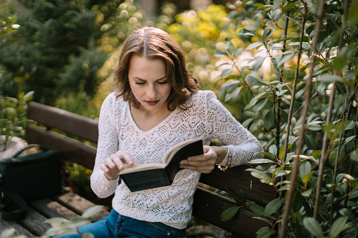 Beautiful young casually dressed woman sitting in the city park and reading an interesting book on a lovely sunny day