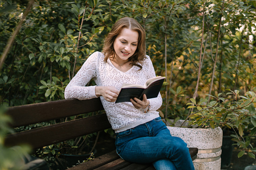 Beautiful young casually dressed woman sitting in the city park and reading an interesting book on a lovely sunny day