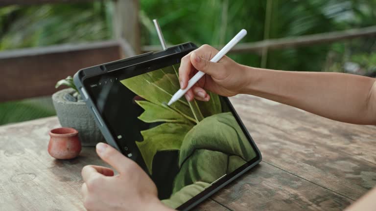 Female artist hands drawing with stylus on tablet at the cafe on tropical island, palm trees. Young designer works remotely open air at exotic location. Pretty woman editing illustration. Close-up.