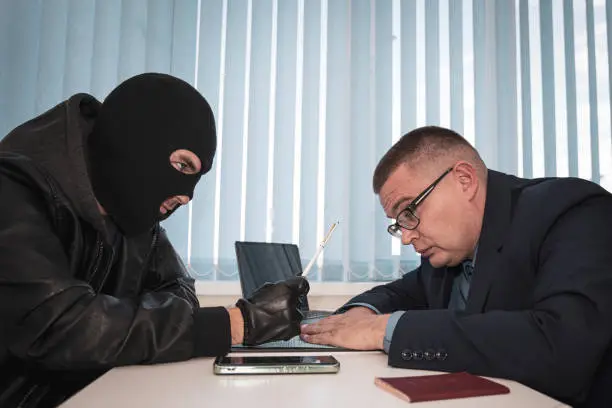Photo of businessman and robbers are sitting at a table. A racketeer in a black balaclava forces to sign a contract. The concept of a raider takeover of the company. dangerous deal