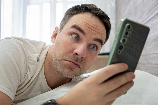Man surprised to hear ringing of alarm smart phone, lies in white bed. Wake up shocked guy late for work in morning. Surprise from the message on smarfton