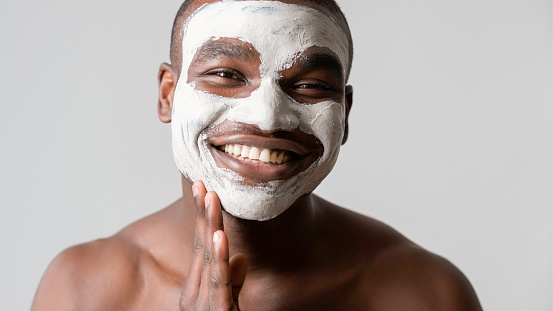 Man cosmetic. Facial mask. Confident satisfied guy enjoying applying face skin pore cleaning white clay smiling isolated on gray background.
