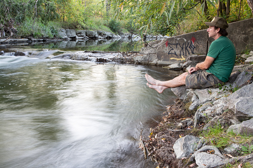 Man in green shirt and green hat with camouflage shorts sitting by the river bed enjoying nature.