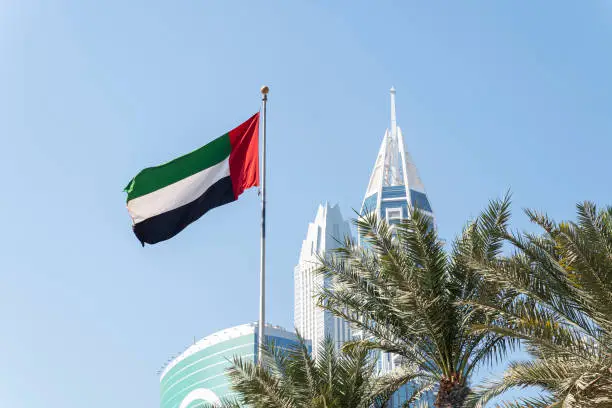Photo of the flag of the UAE on a blue sky background. Flag waving at the top of the modern building, national symbol of United Arab Emirates. National Day and UAE flag day concept.