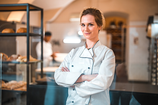 Portrait of a Cheerful Caucasian female shop assistant in an artisan bakery. Standing at a counter with arms crossed looking at camera.