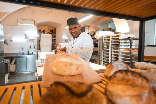 Mature Hispanic baker taking out a freshly baked bread out of the oven. He is using a shovel and putting it on a rack.He enjoys working in an artisan bakery.