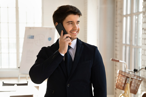 Smiling young Caucasian businessman in formal suit look in distance talk on smartphone gadget in office, happy male boss or CEO have cellphone call speak on modern device, new technology, 5g concept